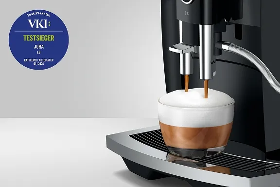 Detailansicht der schwarzen Jura E6, die Milchschaum in ein Cappuccino-Glas füllt. Mit VKI-Testsieger-Siegel.